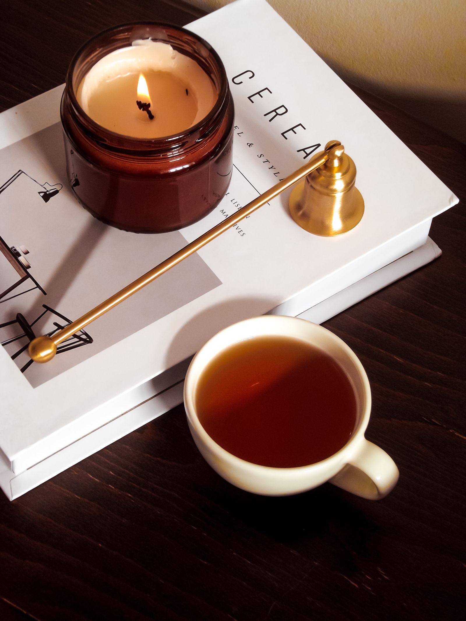 A burning candle beside a book and a cup of tea on a wooden table