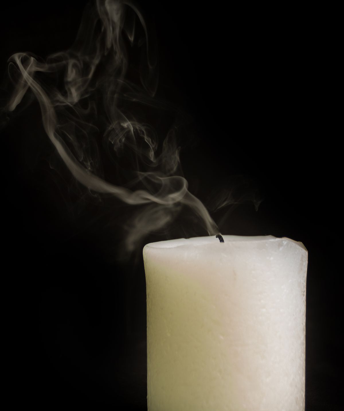 Thin smoke trailing from a white pillar candle's wick, the visible byproduct candle owners ask about as soot