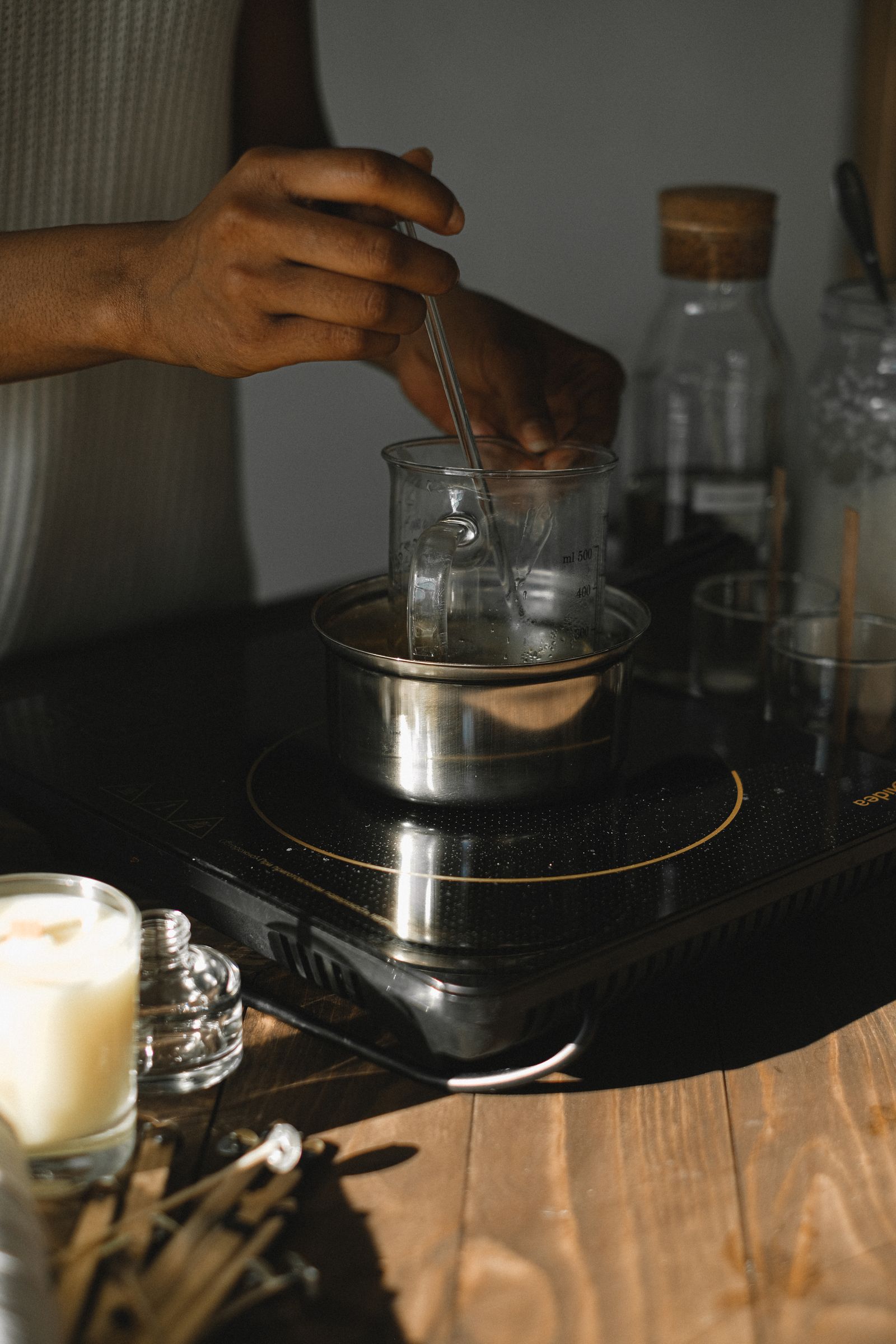 A person stirring melted candle wax in a glass beaker set inside a metal double boiler on an induction stovetop