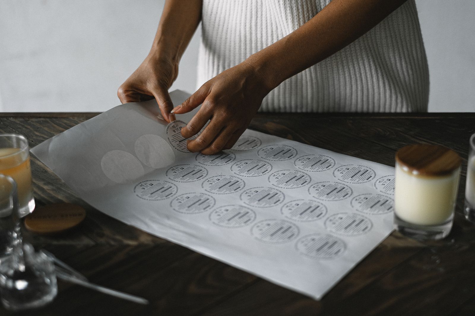 Hands peeling a printed sticker sheet used for labeling handmade candle jars, with a finished wood-topped candle nearby