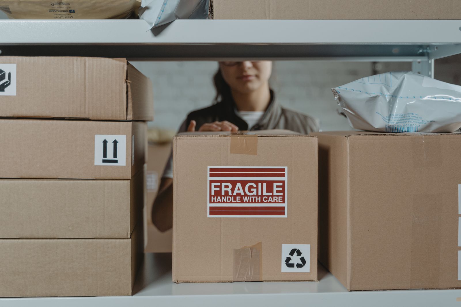 Stacked cardboard shipping boxes on warehouse shelving, one marked FRAGILE — HANDLE WITH CARE, with a person visible working behind them