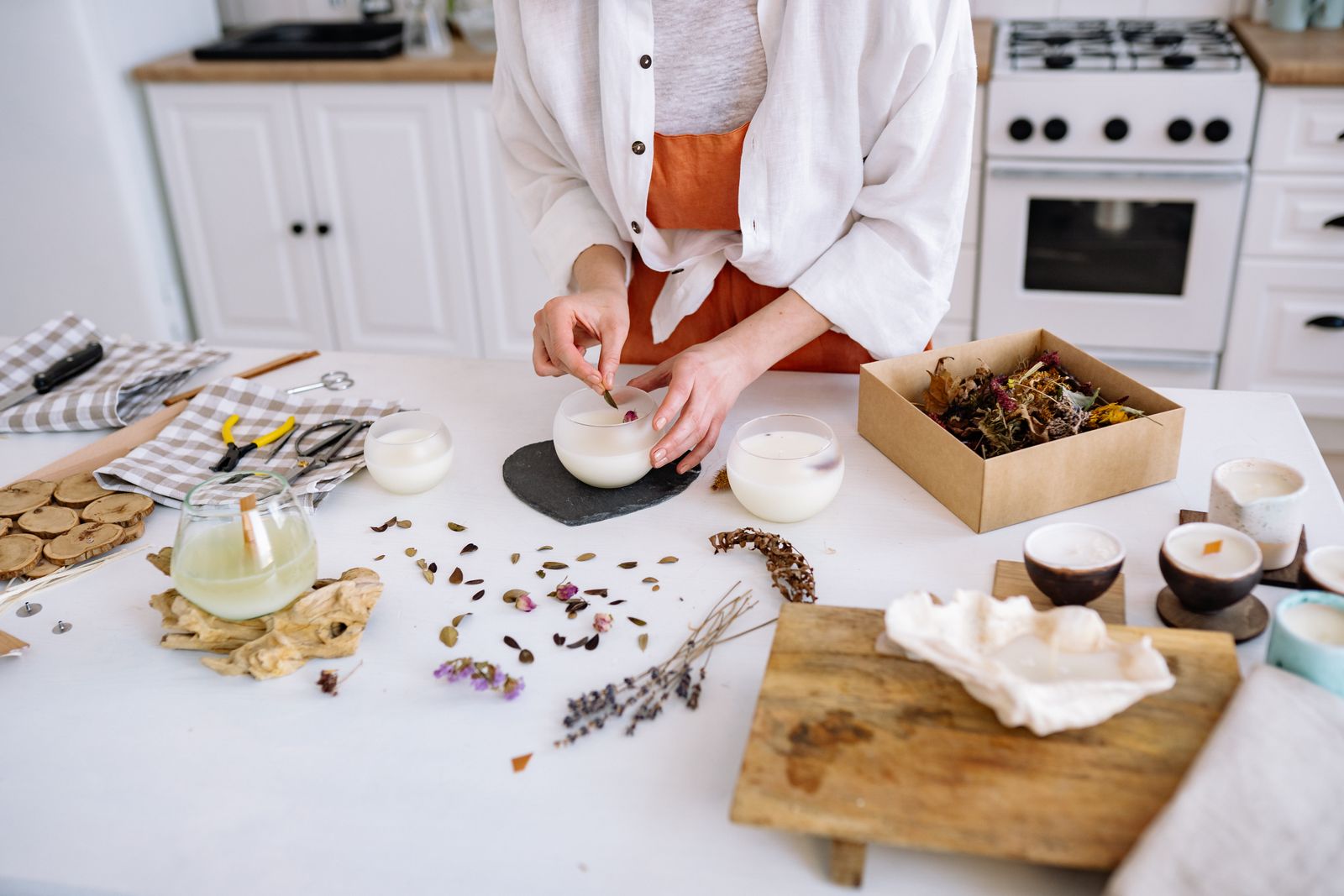 A maker's hands placing a dried flower into a small soy candle on a workshop table surrounded by unpoured jars and dried botanicals